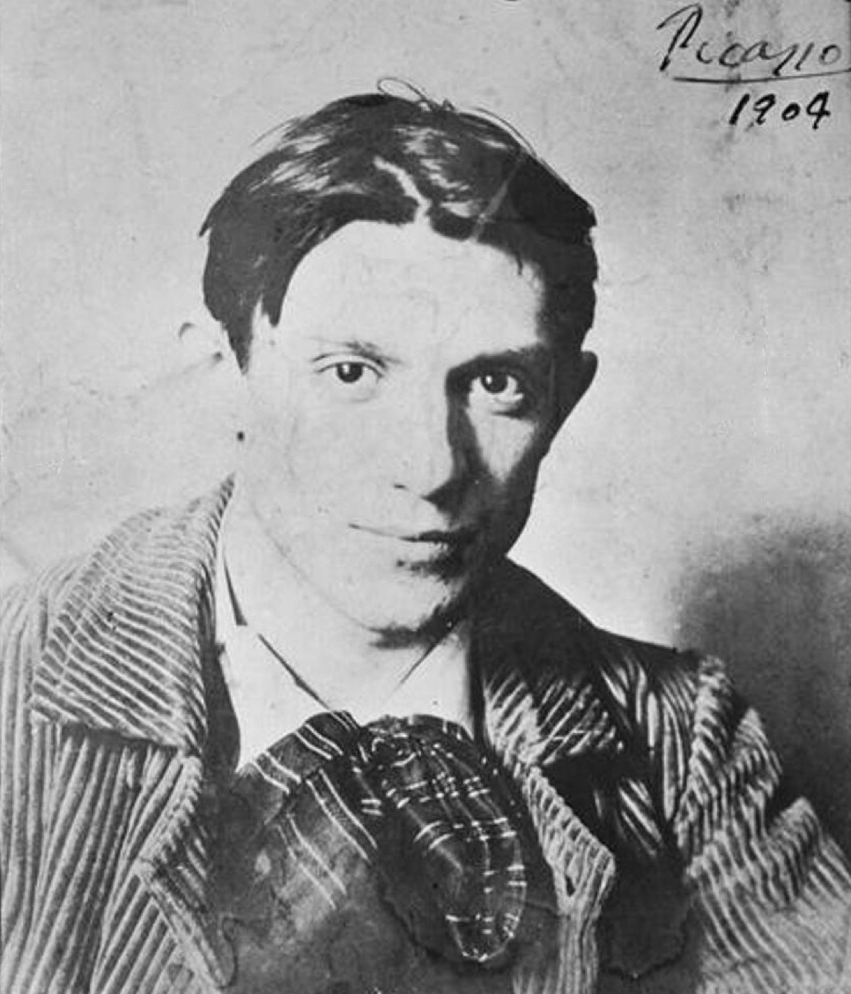 A black-and-white 1904 photographic portrait of a young Pablo Picasso in a corduroy jacket and scarf, signed 'Picasso 1904'.